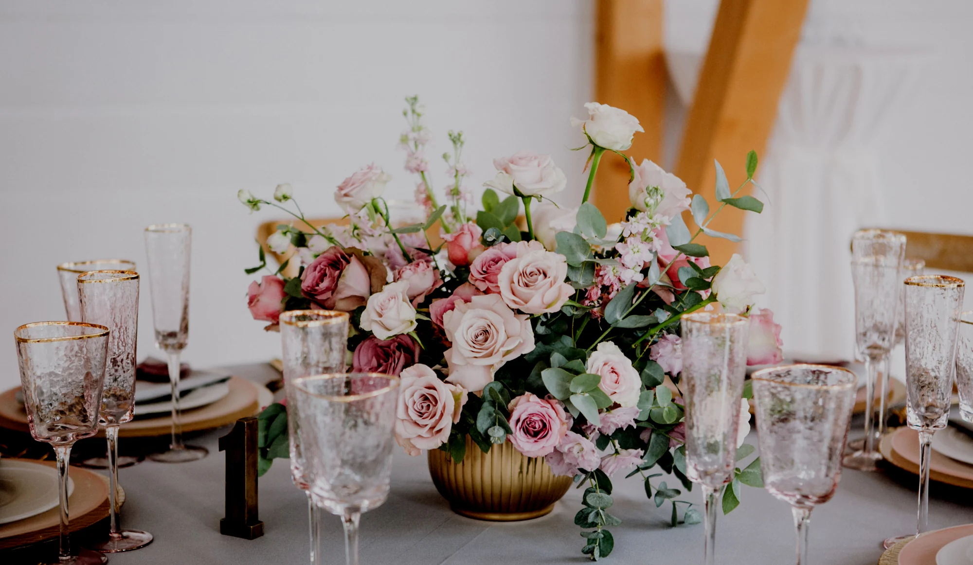 Low centrepiece of blush and dusky pink roses, stocks and eucalyptus in a gold bowl, set among gold rimmed glassware on a wedding table