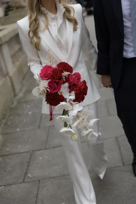 Bride in a white trouser suit carrying a cascading bouquet of red roses and white orchids